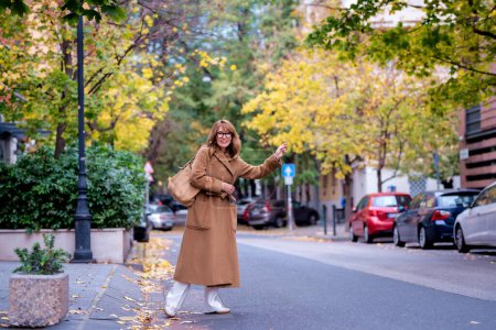 Mujer de mediana edad esperando un taxi al aire libre. Alegre dama moderna con el gesto de bolsa para dar señal al taxista. Longitud completa.