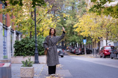 Mujer de mediana edad esperando un taxi al aire libre. Alegre dama moderna con el gesto de bolsa para dar señal al taxista. Longitud completa.