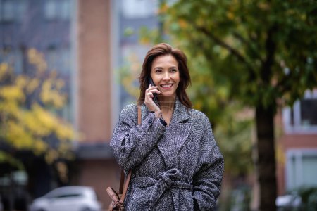 Mid adult woman using smartphone and walking outdoors in city street on autumn day. Attractive female wearing tweed coat and having a call. 