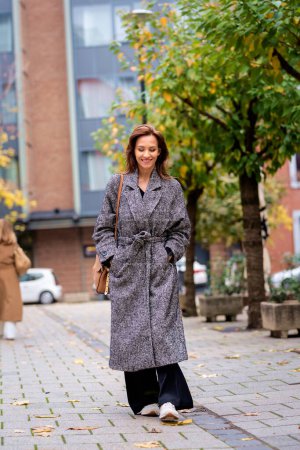 Full length of a mid adult woman with brunette hair walking outdoors in city street on autumn day. She is wearing tweed coat.