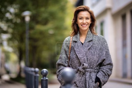 Portrait of a mid adult woman with brunette hair walking outdoors in city street on autumn day. She is wearing tweed coat.
