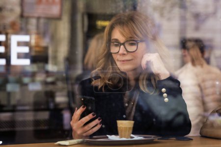 Mujer de pelo rubio sonriente sentada en la cafetería y usando su teléfono inteligente. Atractiva hembra bebiendo café y fotografiada a través de la ventana. 
