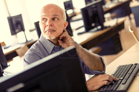 man at the computer listens to a colleague's opinion; he has one hand resting on the keyboard and is in a large room equipped with lots of computers. Open spaces are not conducive to concentration but