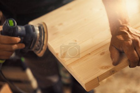The carpenter's hands grip a sander. The wood artisan, an experienced professional, holds a board or piece of wood with strong and precise hands, carefully sanding it with a machine that vibrates sand