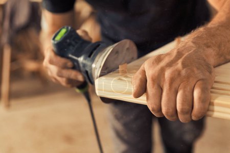 The carpenter's hands grip a sander. The wood artisan, an experienced professional, holds a board or piece of wood with strong and precise hands, carefully sanding it with a machine that vibrates sand
