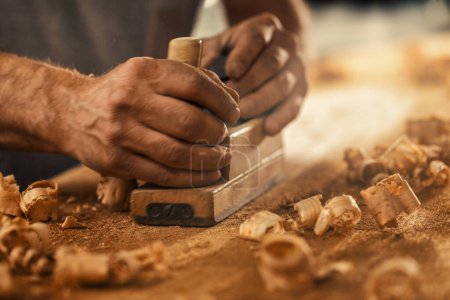 The wood artisan, a robust and strong man known as a carpenter, hand planes a board in the traditional manner, producing shavings of sawdust that look like golden curls of wood. It is a strenuous and 