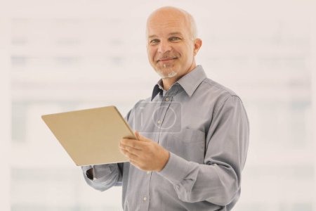 Cheering mature man consults a digital tablet in a big, bright company: with new technology he can talk to an Ai, work remotely, manage projects and teams and control the results