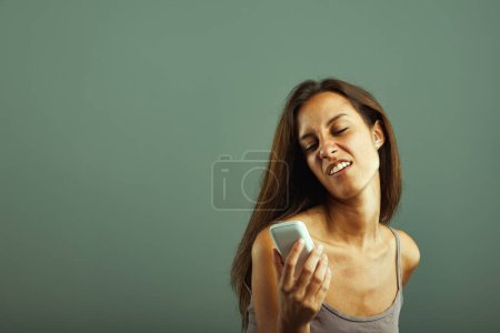 Emotional portrait of young woman, annoyed, mocking phone. Mimics person she dislikes. Envious, impulsive. Brown hair, slim, beautiful, beige tank top. State of mind concept
