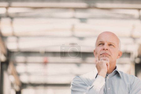 Thinking man on modern metallic background, in sunny open place, touches his chin with his finger. he is bald and has a goatee.