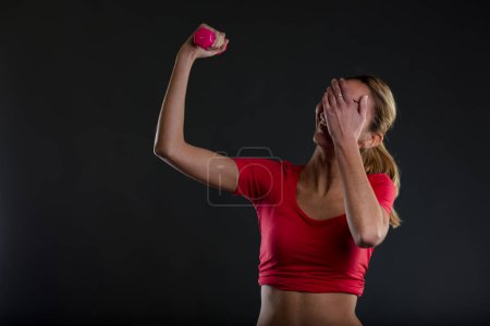 A young, self-ironic woman in a gym, lifts a small pink weight, laughing behind her hand. Wearing a red top, she embraces her responsibility for her fitness without fanaticism