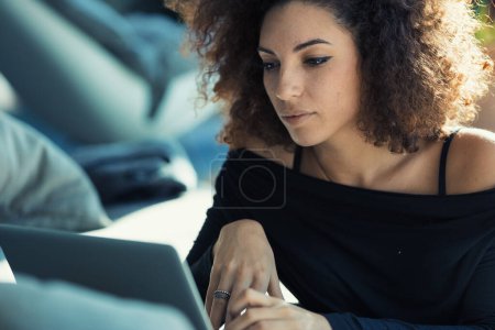 Hermosa mujer rizada, intensa, enfocada en la pantalla de su laptop. Su vestido revela sus hombros seductores mientras se reclina en la alfombra junto al sofá. La luz solar se filtra a través de ventanas anchas, il