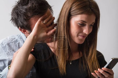Eine Frau benutzt ihr Smartphone, während ihr Mann versucht, von hinten zu gucken. Sie hebt die Hand, um ihm die Sicht zu versperren, und macht ihr Recht auf Privatsphäre geltend. Die Dynamik der privaten Telefonkommunikation hat sich stark verändert.