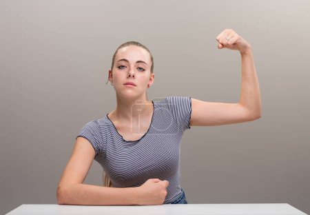 Strong young woman leans on her forearm in a manly way and flexes her other muscle, pulling it with her fist. Her cocky and confident expression says she won't be ordered around, she's her own boss