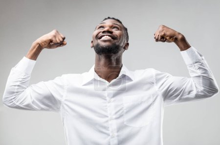 Schwarzer Mann jubelt, die Arme erhoben, lächelnd nach Erfolg durch Anstrengung, nicht nach Glück. Gute Nachrichten feiern, strahlende Zukunft. Ein elegantes weißes Hemd wie ein Geschäftsmann
