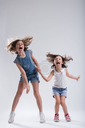In jeans and sneakers, two girls joyfully leap and dance, hands intertwined, tongues out. Their vivacious energy and pure joy hint at a close bond, be it friendship or sisterhood