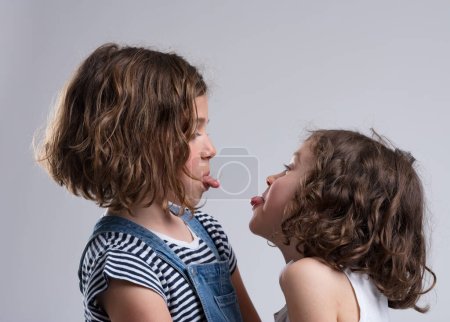 Two young girls, aged 5-7, teasingly stick out their tongues. Likely friends or sisters, they face each other making faces, dressed casually. They laugh joyfully