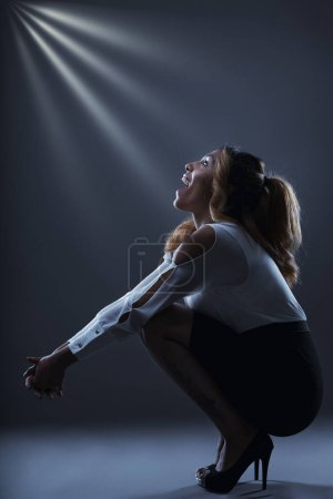 Woman in heels sits, basking in a spotlight of triumph, her joy unmistakable in elegant attire