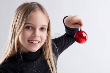 cheerful young girl holds a red Christmas ornament, her smile reflecting festive joy