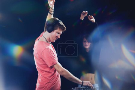 DJ raises hand in joy, woman in background dancing, both encapsulated by stage lights