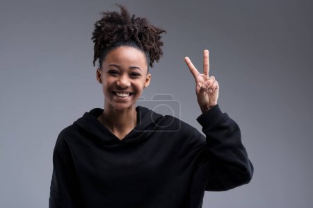 A slim, pretty woman with dark brown skin and afro hair giggles, showing a victory sign. Her cheer and happiness in a black sweatshirt are infectious. She celebrates her success, radiating charm