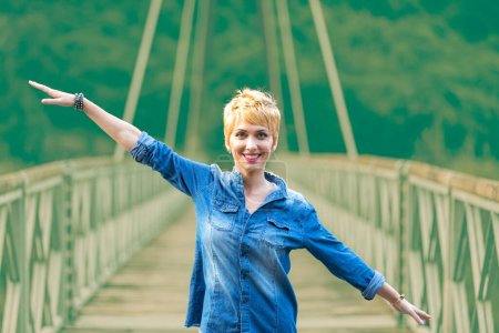 Freude strahlt die Frau in Jeans aus, die Arme weit aufgerissen auf einer Brücke über spiegelndem Wasser