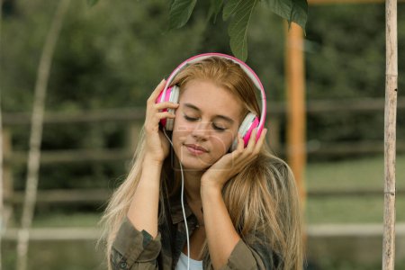 Young woman enjoys music with pink headphones, eyes closed in serene moment, surrounded by nature