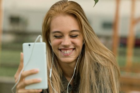 Strahlendes Lächeln auf der jungen Dame, als sie einen Videoanruf genießt, Kopfhörer in der Hand und ein Smartphone