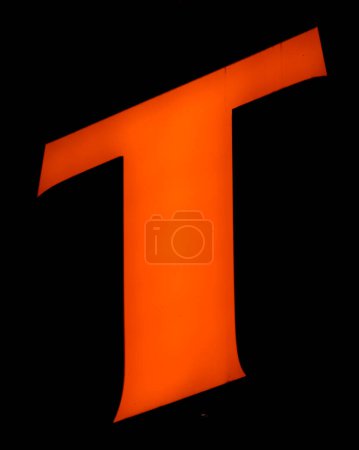 Captured from below is a striking, luminous 'T' set against a dark backdrop. Its audacious orange hue stands out vividly, contrasting sharply with its surroundings. Such a design choice might signify 