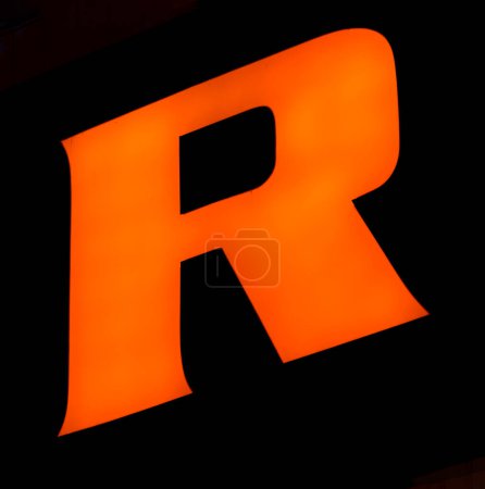 Von unten eingefangen ist ein markantes, leuchtendes "R" vor dunklem Hintergrund. Sein kühner orangefarbener Farbton sticht lebhaft hervor und steht in scharfem Kontrast zur Umgebung. Eine solche Gestaltungswahl könnte bedeuten, dass 