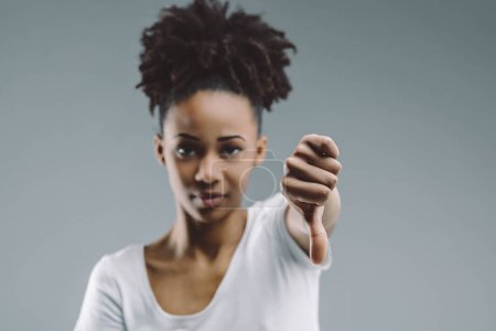 Mujer afro-peluda da gesto de pulgares hacia abajo, con una mirada perspicaz, sobre un fondo liso