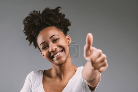 Mujer joven radiante en blanco, su pelo afro y sonrisa radiante complementando sus entusiastas pulgares hacia arriba