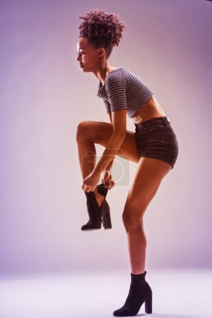 Jeune femme avant-gardiste en chemise rayée et short met sa botte, mettant en valeur la grâce et le style