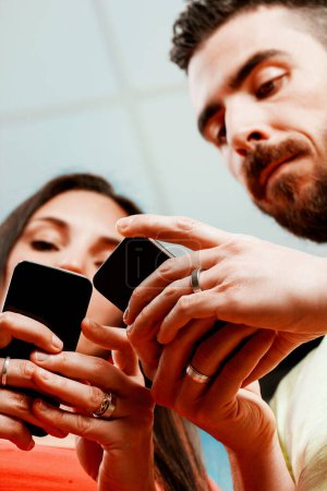 Focused young man and woman, both wearing rings, engage with their own smartphones in a close, personal space