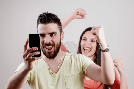 Celebrando un logro significativo, una pareja captura alegremente la victoria con un teléfono inteligente, con los puños apretados en la emoción