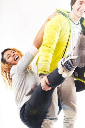Jeune homme vêtu d'une veste jaune et bonnet soulève une jeune femme riante dans un pull gris décontracté, profitant tous deux d'un moment ludique et joyeux ensemble