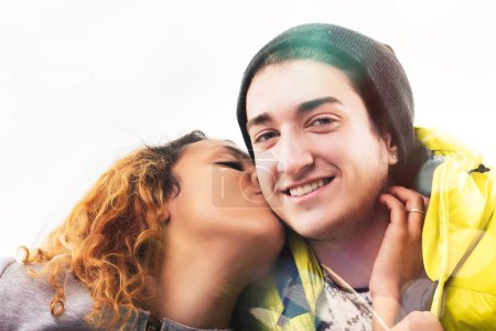 young woman with curly hair affectionately kisses a smiling young man on the cheek, both dressed in warm, casual winter attire