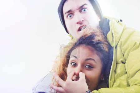 Humorous and candid scene with a young man in a beanie and yellow jacket playfully covering a woman's mouth, both showing playful expressions