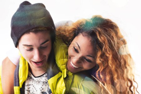Young woman with curly hair wearing a bright yellow jacket embraces a young man in a dark beanie and vest, both smiling in a candid, joyful moment