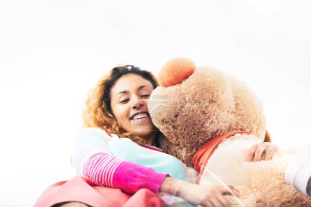 Strahlende junge Frau umarmt einen großen Teddybär mit einem fröhlichen Lächeln, ihr buntes Gewand trägt zur fröhlichen Szene bei