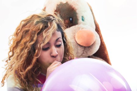 Young woman inflates a large pink balloon, with a stuffed dog toy looking over her shoulder, expressing a playful mood