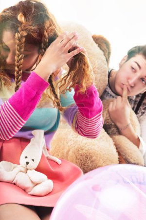 Light-hearted interaction between a curly-haired woman and a young man, both engaging with playful props and a big teddy bear