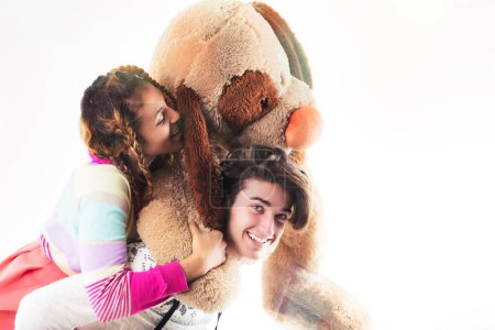 Two friends or siblings in a playful setting with a giant teddy bear, one wearing a furry hat and the other a crown