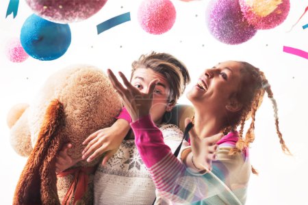Junge Männer und Frauen, möglicherweise Geschwister oder Freunde, beschäftigen sich spielerisch mit einem Stoffbär und Luftballons und tragen skurrile Hüte