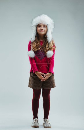 cheerful young girl in a festive white hat and bright pink sweater, paired with patterned brown shorts, poses playfully