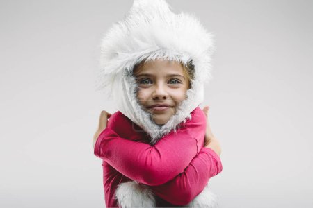 Young girl in a whimsical white winter hat hugs herself warmly, her joyful eyes and gentle smile conveying comfort and happiness