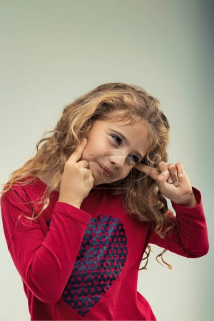 Imaginative young girl with curly blonde hair gestures to her head, wearing a red shirt with a heart pattern, suggesting creativity