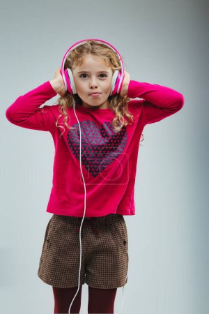 Blondes junges Mädchen in rosafarbenem Pullover und dekorativer Shorts hält Kopfhörer in der Hand, Lippen in verspieltem Ausdruck, vor grauem Hintergrund