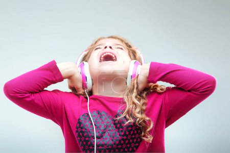 Joyous young girl with curly blonde hair, dressed in pink, uses white headphones, singing passionately with a beaming expression