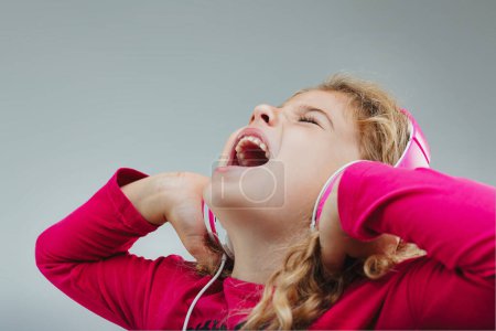 Junges Mädchen in leuchtendem rosa Top, genießt Musik durch weiße Kopfhörer, singt fröhlich mit geschlossenen Augen, die Hände vor dem Gesicht