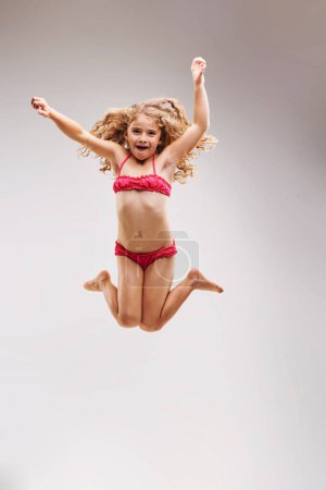 La niña en traje de baño rosa salta tanto que parece estar volando por el aire, con su pelo rubio rizado a su alrededor. Orgulloso y fuerte.